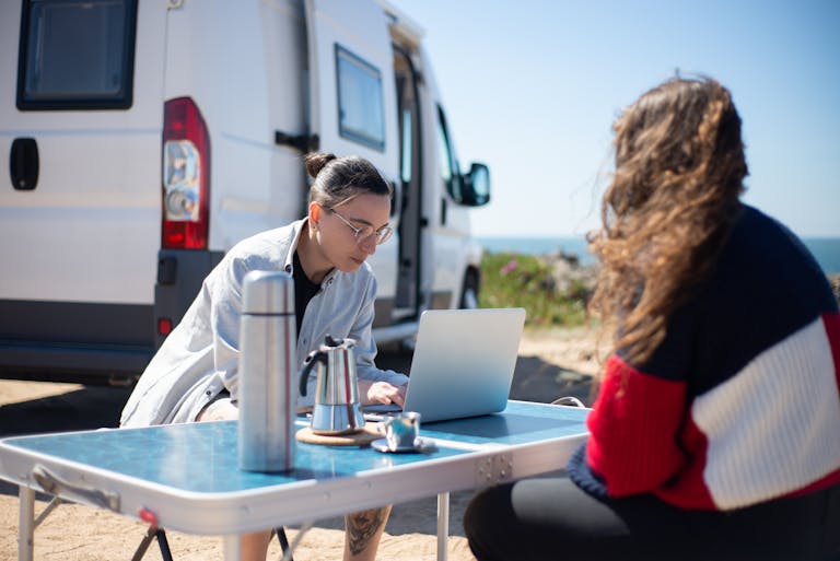 Two adults working on a laptop by a camper van at a scenic beach location in Portugal.